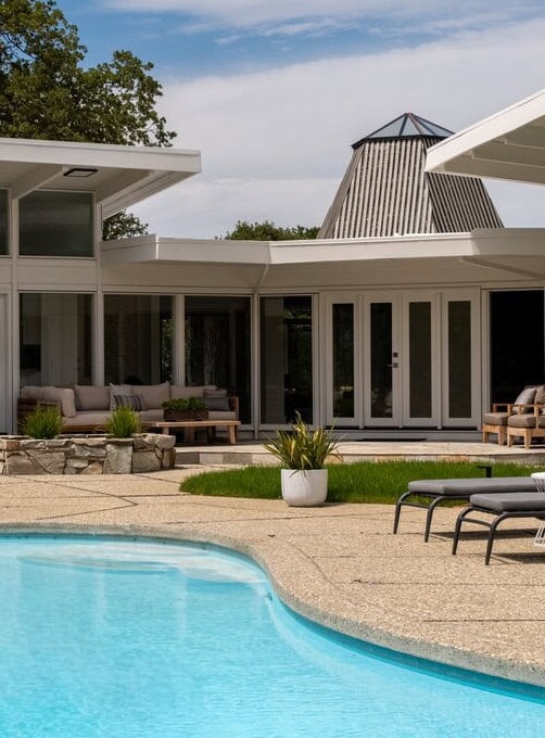 Modern Home Courtyard With A Clear Blue Swimming Pool, Lounge Chairs, Outdoor Dining Table, And Green Landscaping On A Sunny Day. White Walls And Large Glass Windows Surround The Space.