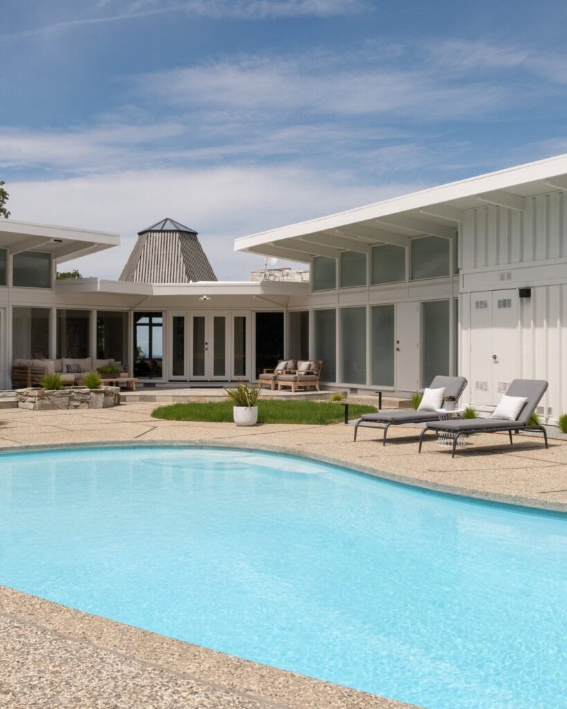 A Modern White House With Large Windows Surrounds A Backyard Patio Featuring Lounge Chairs, Potted Plants, Outdoor Seating, And A Clear Blue Swimming Pool Under A Sunny Sky.