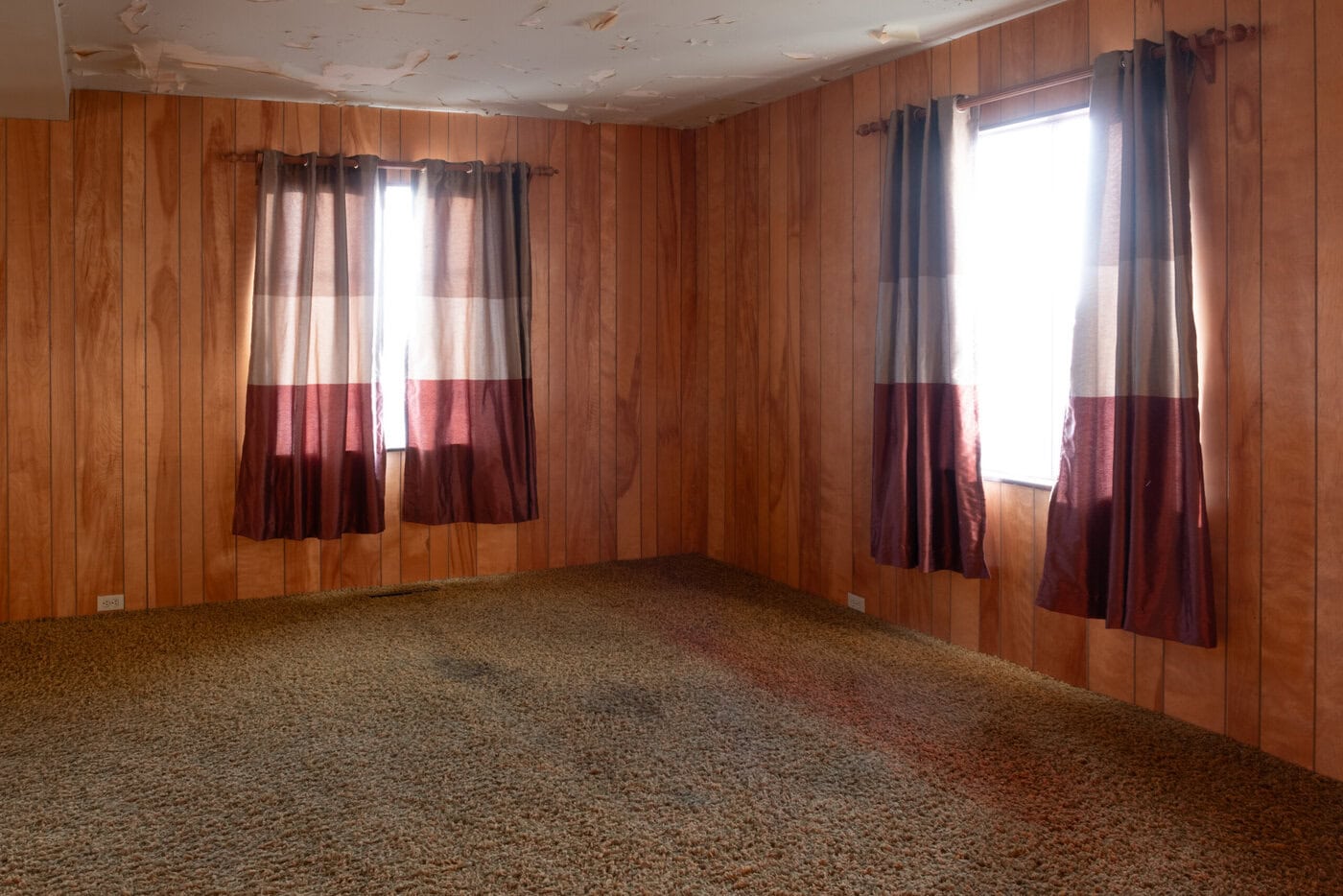 An Empty San Francisco Home With Wood-Paneled Walls, Two Windows Adorned With Red And White Curtains, A Brown Carpet Showing Visible Stains, And A Ceiling With Peeling Paint.