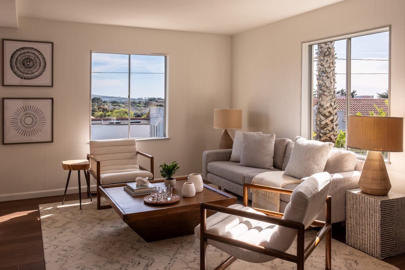 A Bright Living Room With Large Windows, Beige Sofa, Two White Cushioned Chairs, Wooden Coffee Table, Lamps, Framed Art, And A Neutral Rug—Perfect For Relaxing Or Showcasing Your Home If You’re Ready To Sell Or Upgrade. Sunlight Streams In, Creating A Warm Atmosphere.