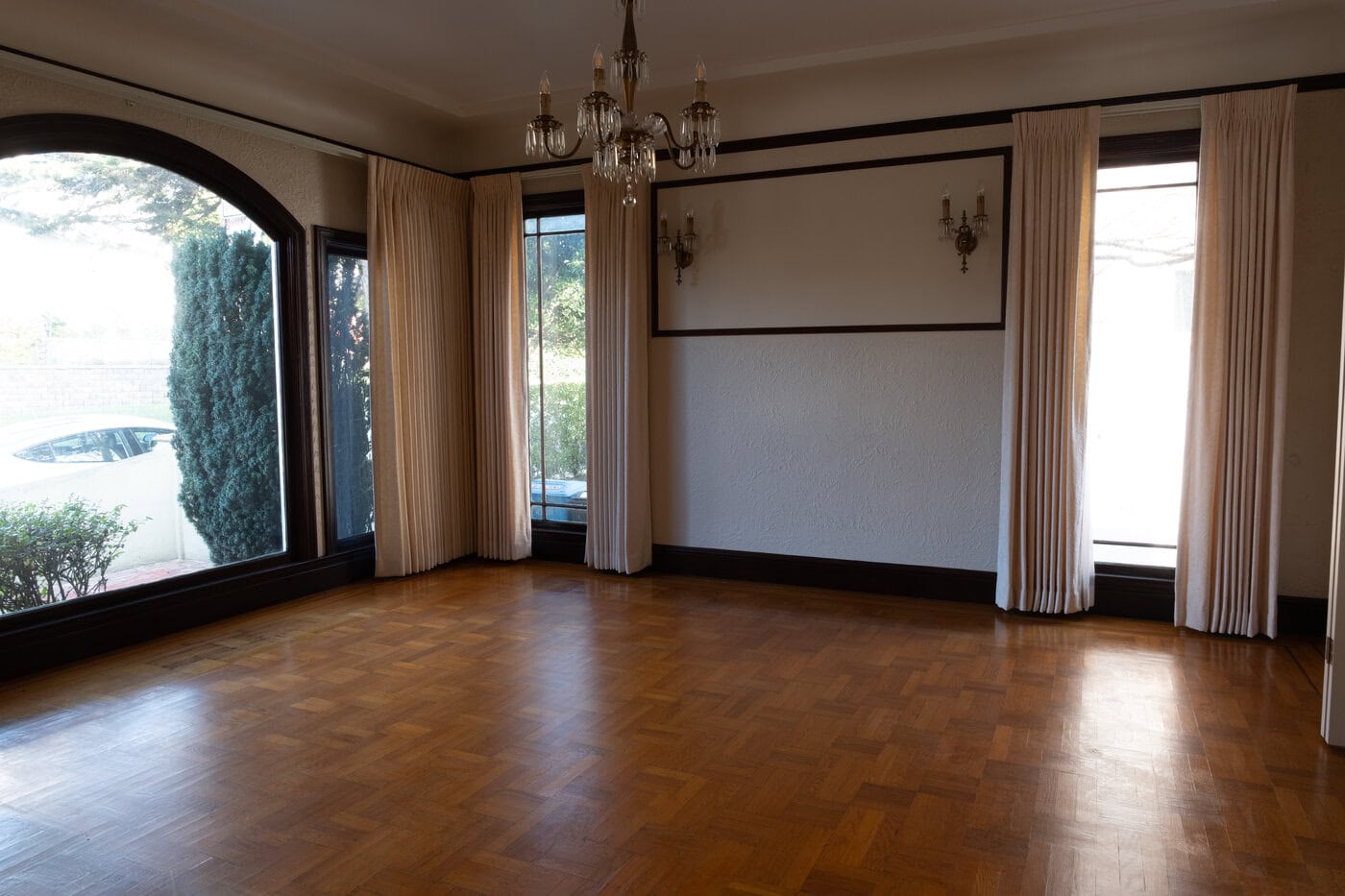 A Spacious, Empty Room In A San Francisco Home Features Parquet Wood Flooring, Large Windows With Cream Curtains, A Chandelier, And Wall Sconces. Sunlight Pours Through The Windows, Illuminating The Floor—Perfect For Your Contractor’s Next Project.