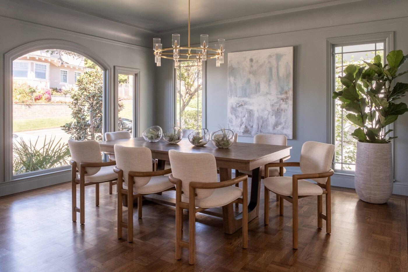Upgrade Your Home With A Modern Dining Room Featuring A Wooden Table, Eight Cream Upholstered Chairs, Glass Terrariums As Centerpieces, Large Windows, A Potted Plant, Abstract Wall Art, And A Contemporary Chandelier.