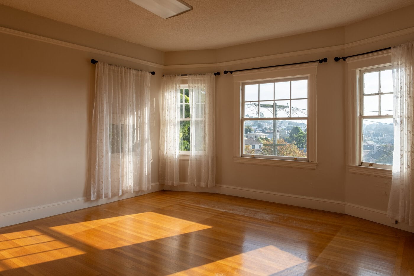 A Bright, Empty Room With Hardwood Floors, Cream-Colored Walls, And Three Large Windows With Sheer White Curtains Letting In Natural Sunlight—A Perfect San Francisco Home Upgrade With Views Of Trees And City Buildings Outside.