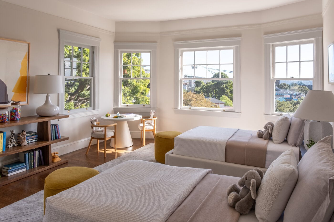 A Bright San Francisco Bedroom With Two Neatly Made Twin Beds, Plush Toys, Yellow Ottomans, A Wooden Shelf With Books And Decor, And Large Windows Letting In Natural Light—Perfect For An Upgrade To Your Home.