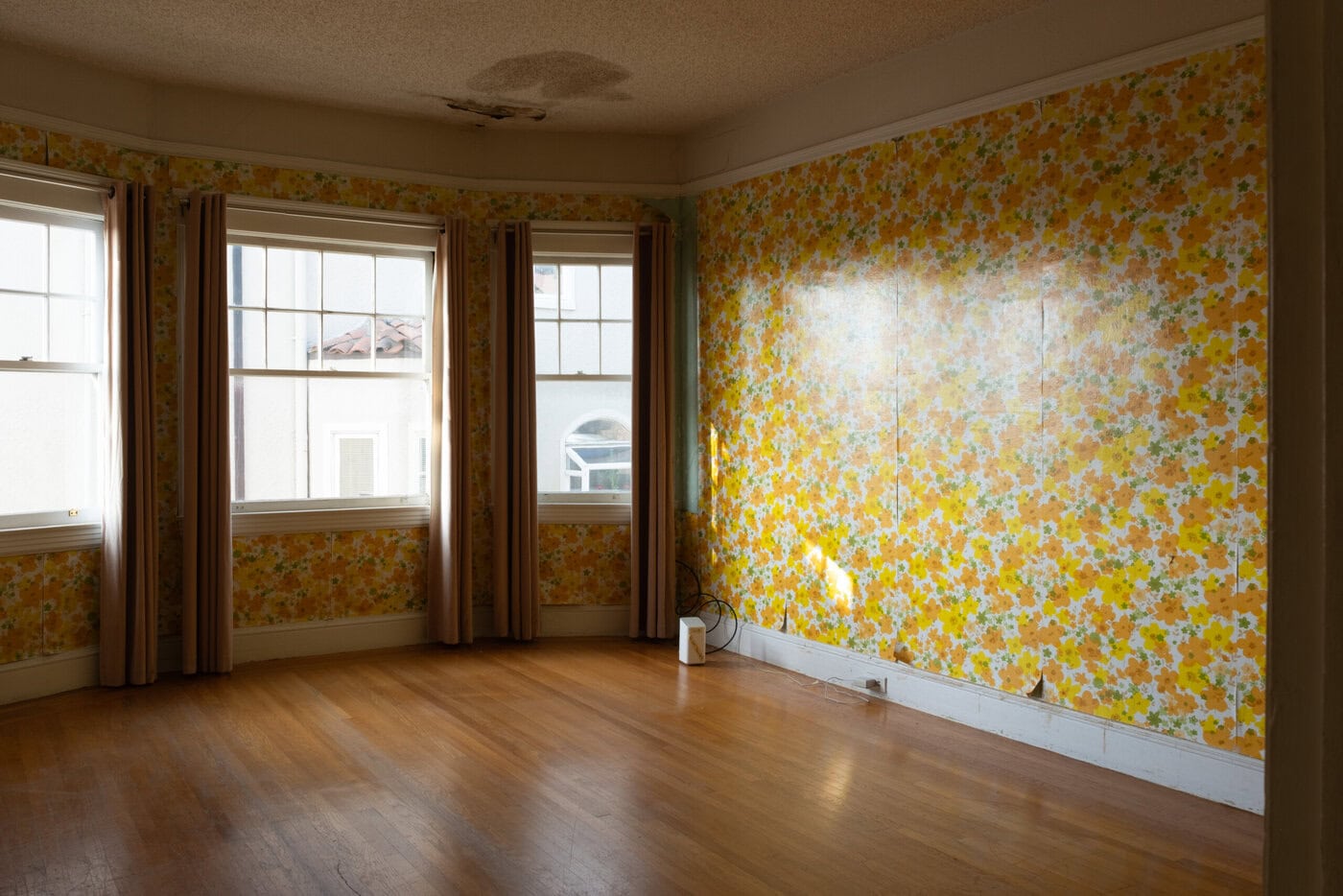 An Empty Home With Wooden Floors, Three Large Windows With Beige Curtains, And Bright Yellow Floral Wallpaper. A Lamp Sits On The Floor Against One Wall, Highlighting Potential To Upgrade Before You Sell. A Water Stain Marks The Ceiling.