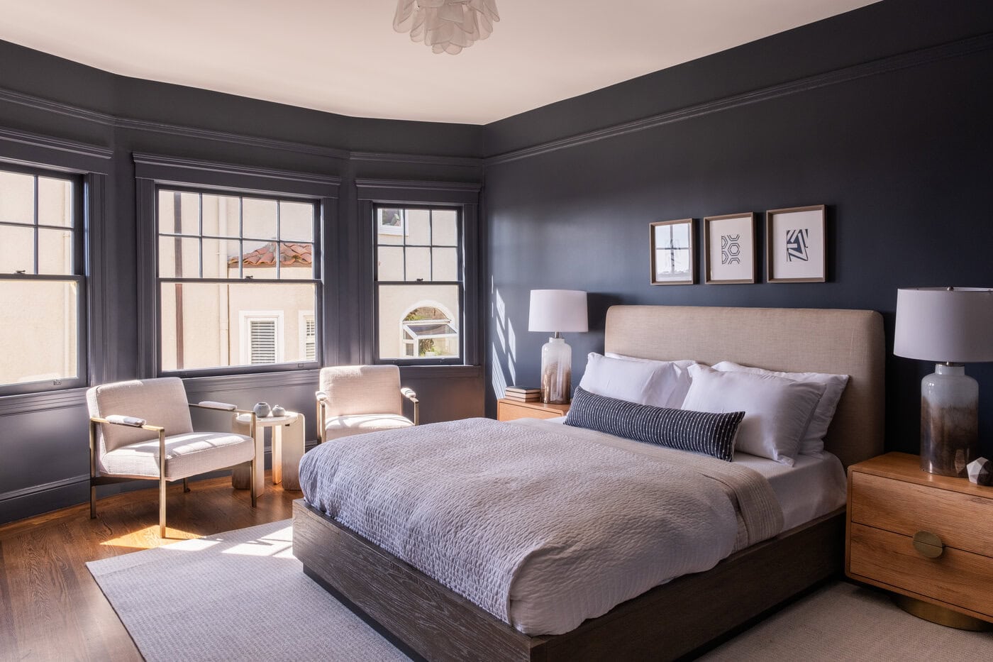A Modern San Francisco Bedroom With Dark Walls, Large Windows, A Beige Upholstered Bed With White Bedding, Wooden Nightstands With Lamps, Two Armchairs By The Window, And Framed Art Above The Bed—A Stylish Home Upgrade Filled With Natural Light.