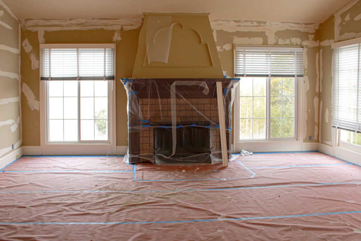 A San Francisco Home Living Room Under Renovation With Drywall Patches On The Walls, A Covered Fireplace, And Protective Plastic And Paper Sheeting On The Floor. Sunlight Streams Through Two Large Windows—Perfect Updates To Help Sell.
