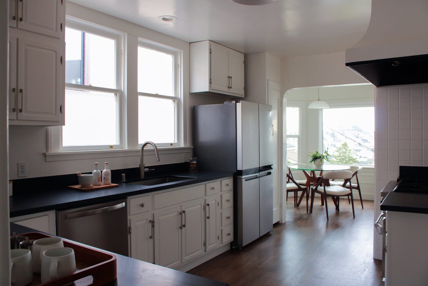 Bright, Modern Kitchen With White Cabinets, Black Countertops, Stainless Steel Appliances, And A Window Above The Sink. This San Francisco Upgrade Features A Dining Area With A Round Table And Chairs By Large Windows.