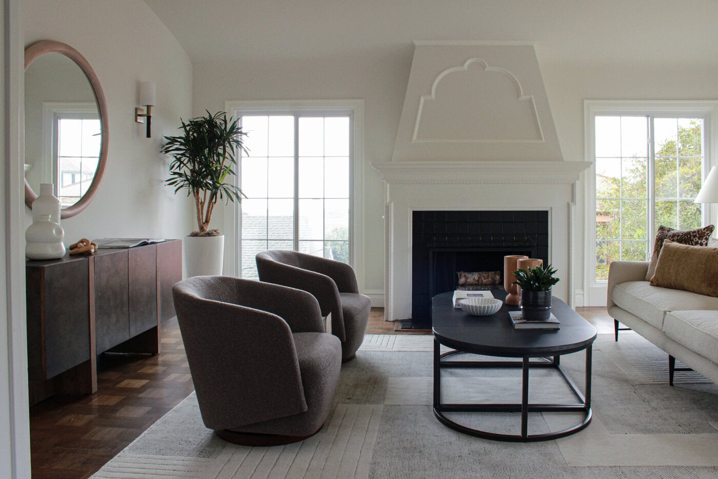 A Modern San Francisco Living Room With Two Gray Swivel Chairs, A Beige Sofa, Black Oval Coffee Table, Fireplace, Large Windows, Round Mirror Above A Console Table, Potted Plant, And Stylish Neutral Decor—Perfect To Call Home Or Sell.