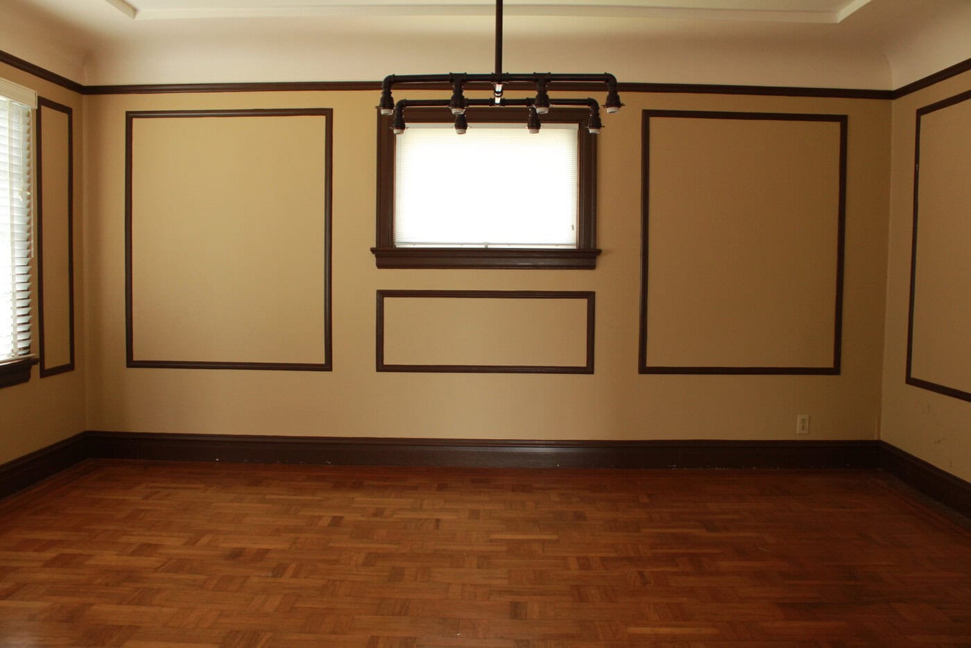 An Empty Room In A San Francisco Home With Tan Walls, Dark Brown Trim, Wood Parquet Flooring, A Small Window With Blinds, And A Ceiling Light Fixture Featuring Exposed Bulbs And A Black Pipe Design—Ready For You To Sell Or Personalize.