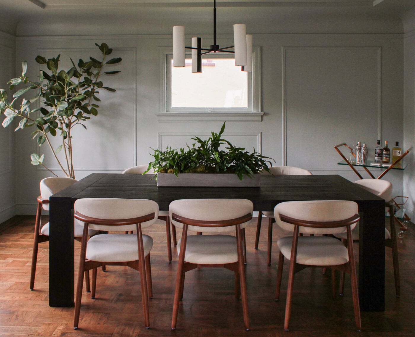 A Modern Dining Room Perfect For Your Home, Featuring A Dark Rectangular Table, Eight Light Cushioned Chairs, A Large Plant Centerpiece, Tall Potted Plant, Window, And A Small Bar Cart With Bottles And Glasses In The Corner.