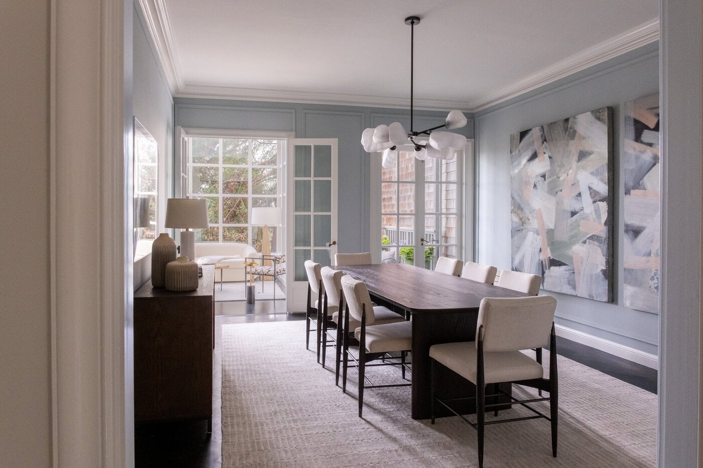 A Modern Dining Room Perfect For Your Home, Featuring A Dark Wooden Table, Eight Cream Chairs, Abstract Art On Light Blue Walls, A Contemporary Light Fixture, Large Windows, And A View Into A Bright Adjacent Room—An Ideal Upgrade For Stylish Living.
