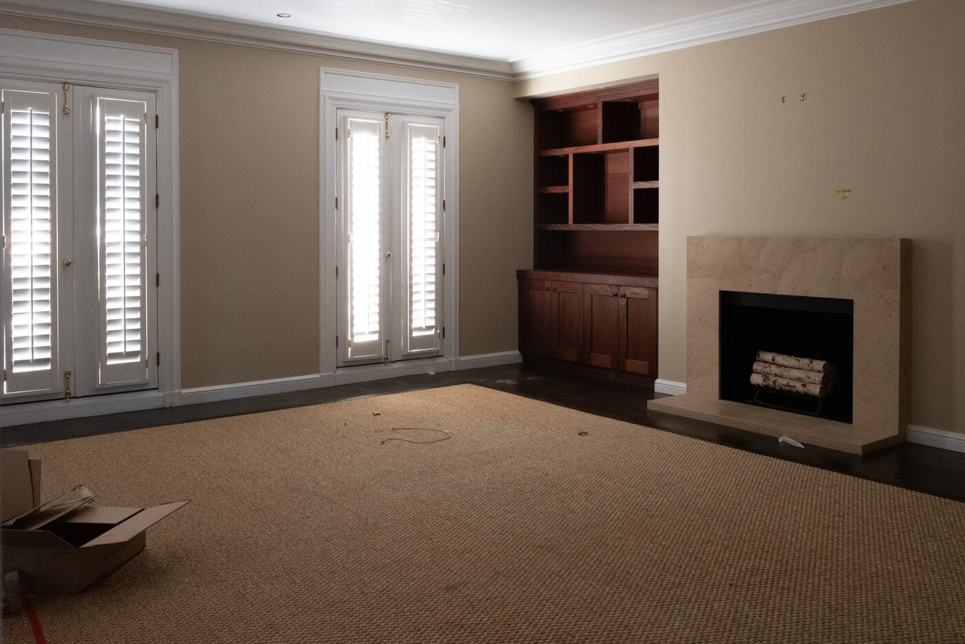 A Mostly Empty Living Room With Beige Walls, A Tan Rug, French Doors With White Shutters, A Fireplace With Logs, Built-In Wooden Shelves, And An Open Cardboard Box Awaits Your Next Home Upgrade In San Francisco.