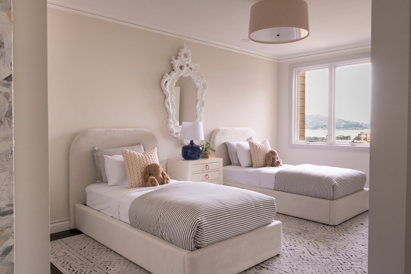 A Bright Bedroom With Two Twin Beds, Each With Striped Bedding And Pillows, Makes A Cozy Retreat. A Nightstand With A Blue Lamp Sits Between The Beds, While A Contractor-Approved Upgrade Includes A Decorative Mirror And Large Window For Scenic Home Views.