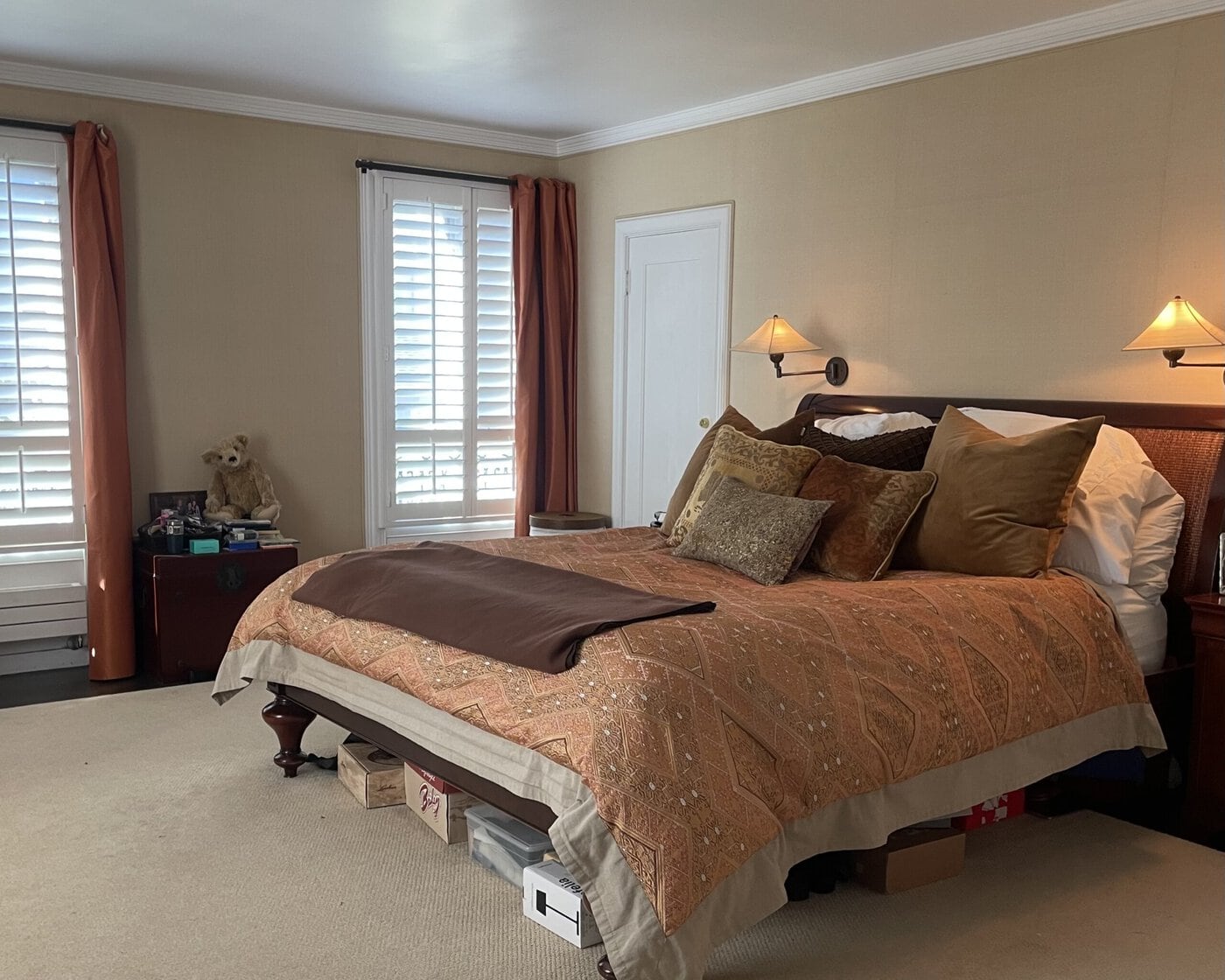 A Neatly Made Bed With Patterned Orange And Brown Bedding Sits Against The Wall In A Cozy Bedroom—Perfect For Anyone Looking To Upgrade Their Home. Pillows, Wall-Mounted Lamps, Storage Boxes, And Windows With Shutters Complete The Inviting Space.