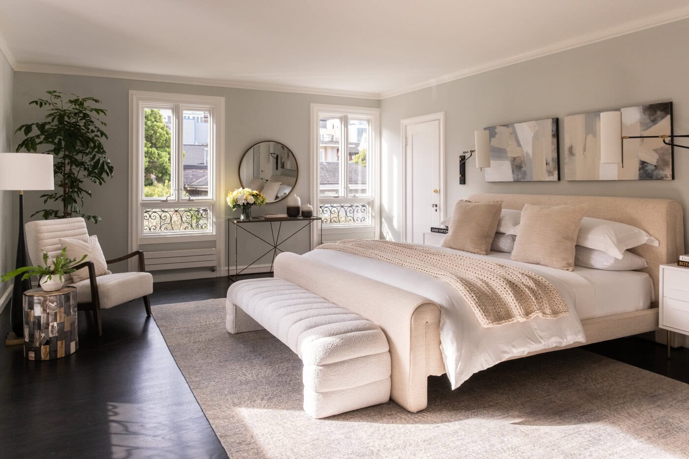 Upgrade Your Home With This Modern, Spacious Bedroom Featuring A Large Bed, Light Upholstered Bench, Abstract Wall Art, Two Windows, A Round Mirror, Plants, And Minimalist Decor In Neutral Tones. Natural Light Fills The Room.