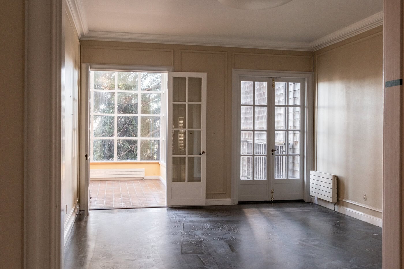 Empty Room With Light Brown Walls, Dark Flooring, And Two Sets Of Glass French Doors. Perfect For A Contractor Or Anyone Looking To Enhance Their Home, The Sunroom Welcomes Natural Light Through Large Windows And A Tiled Floor.
