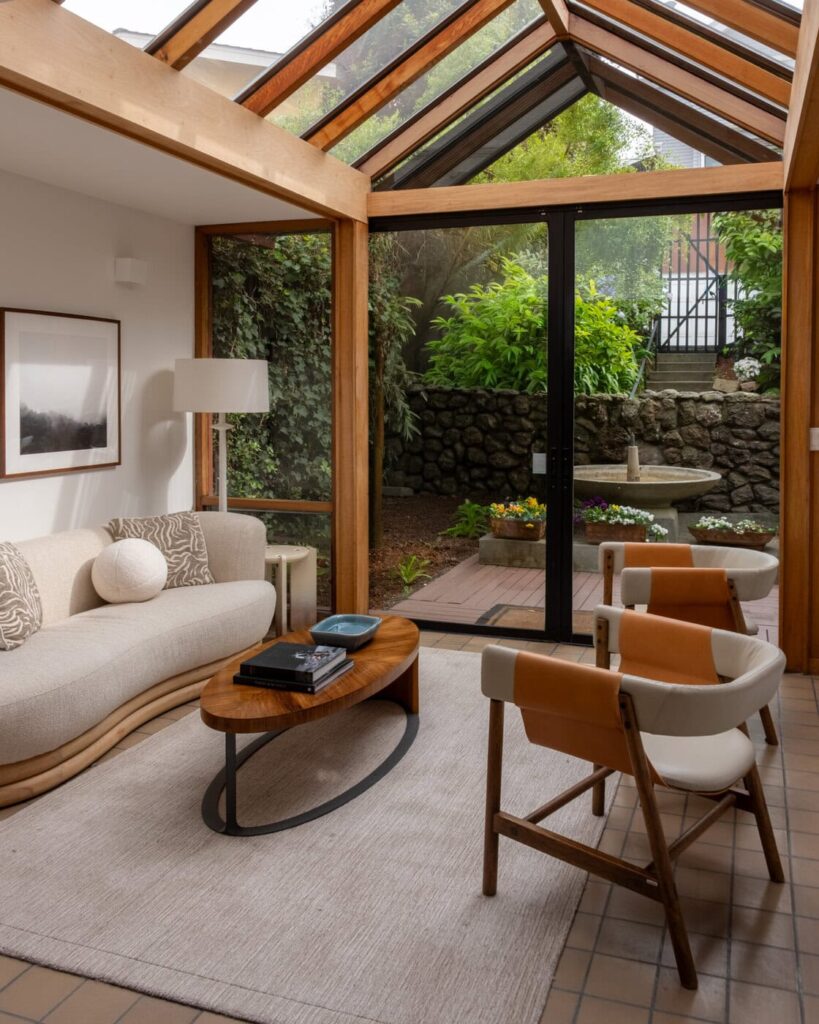 A Cozy Sunroom With A Glass Ceiling, Beige Sofa, Two Leather Chairs, A Wooden Coffee Table, And Sliding Glass Doors Opening To A Garden With Lush Greenery, Flowers, And A Stone Fountain.