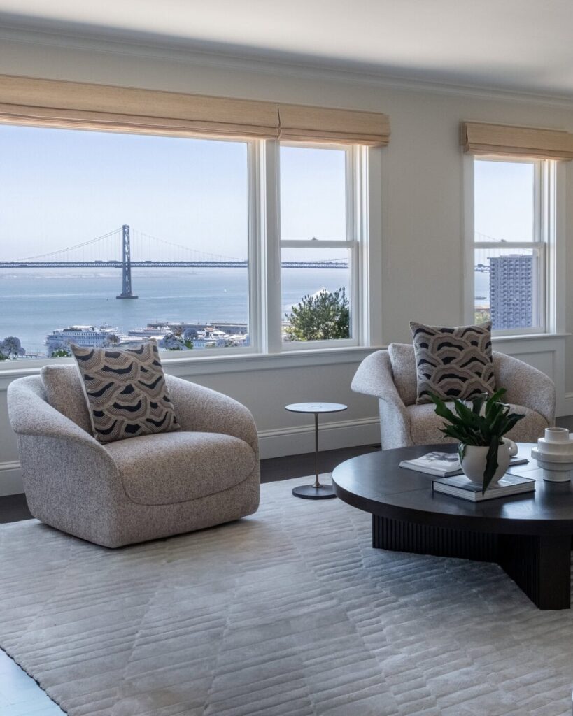 Two Modern Armchairs With Patterned Pillows Sit By Large Windows Overlooking A Bay And A Suspension Bridge. A Round Coffee Table With Decor And A Plant Is Placed On A Light-Colored Rug In The Bright, Airy Room.