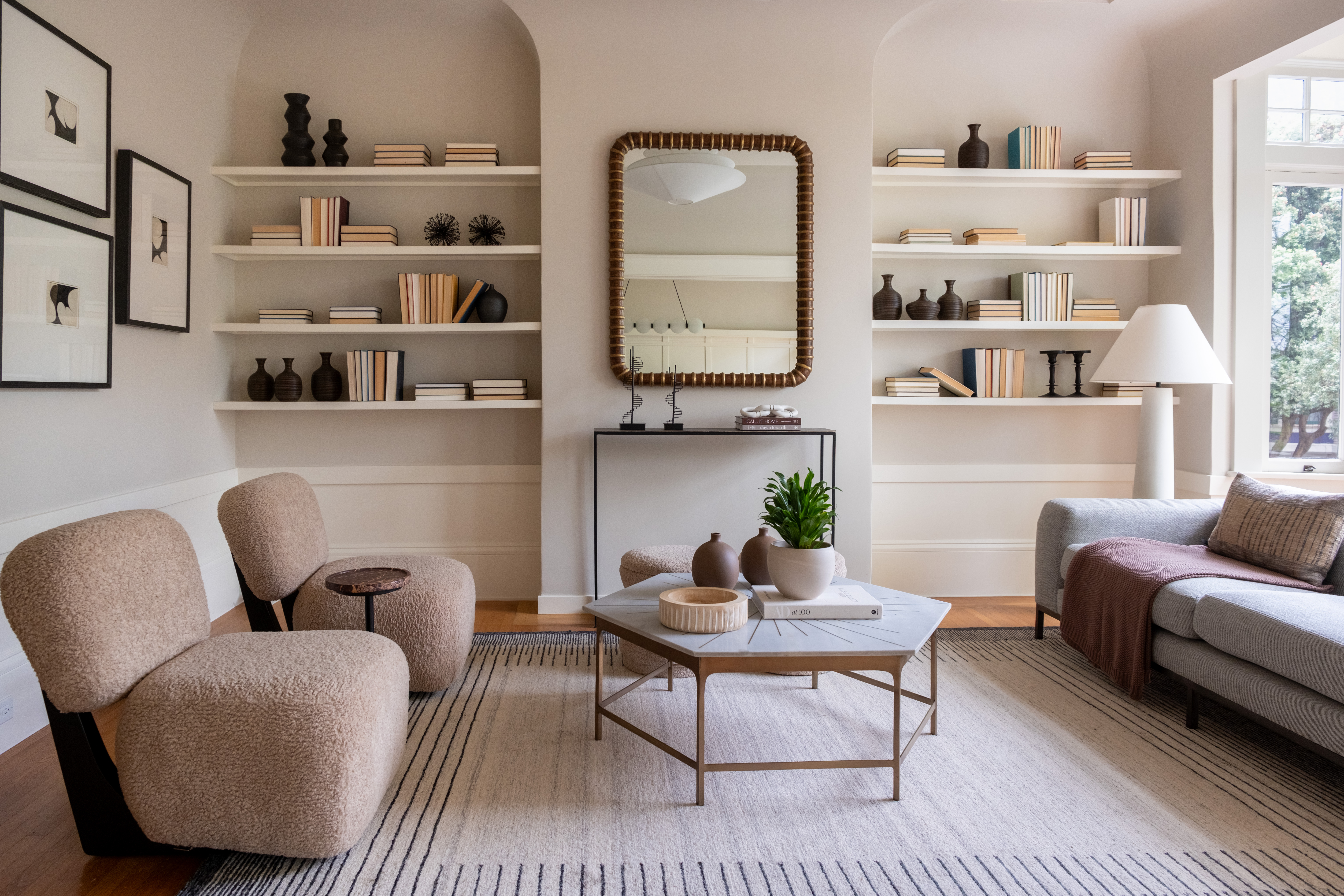 Modern Living Room With Neutral Tones, Two Beige Chairs, A Gray Sofa, A Striped Rug, A Coffee Table With Decor, Wall Shelves With Books And Vases, A Large Mirror, And A Floor Lamp By A Window.