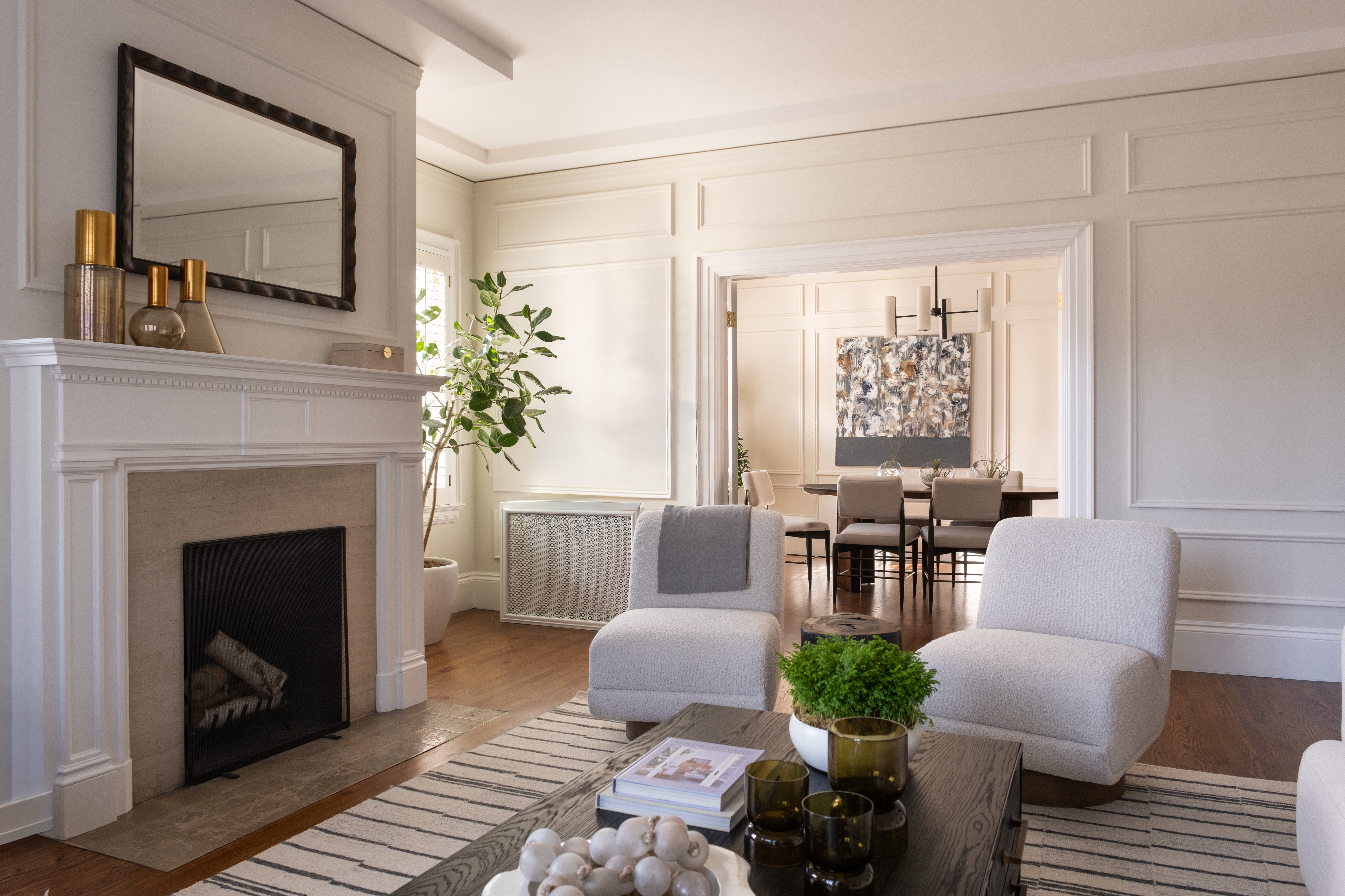 Bright Living Room With White Walls, Modern Furniture, A Striped Rug, A Fireplace With A Mirror Above It, And A Coffee Table With Decor. Dining Area Is Visible In The Background.