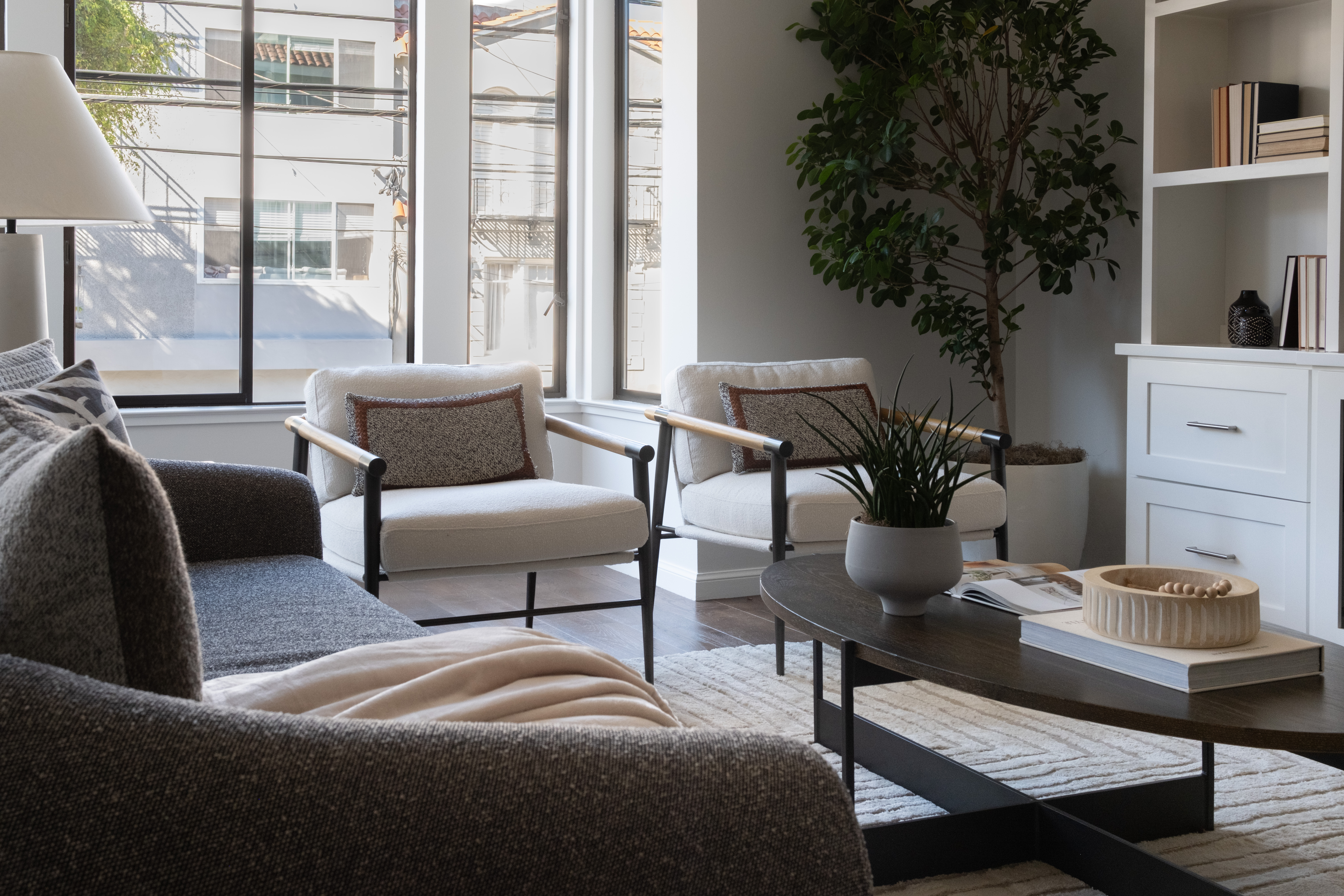 Modern Living Room With Gray Sofa, Two White Armchairs, A Round Coffee Table, Potted Plants, And Built-In Shelving. Large Windows Let In Natural Light, Creating A Cozy And Inviting Atmosphere.