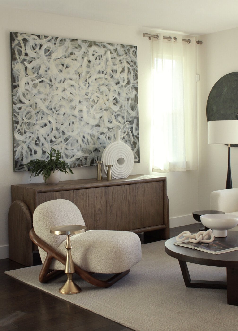 Modern Living Room With A Unique Armchair, Round Side Table, Wooden Credenza, Abstract Wall Art, Neutral Decor, And Sunlight Streaming Through White Curtains.
