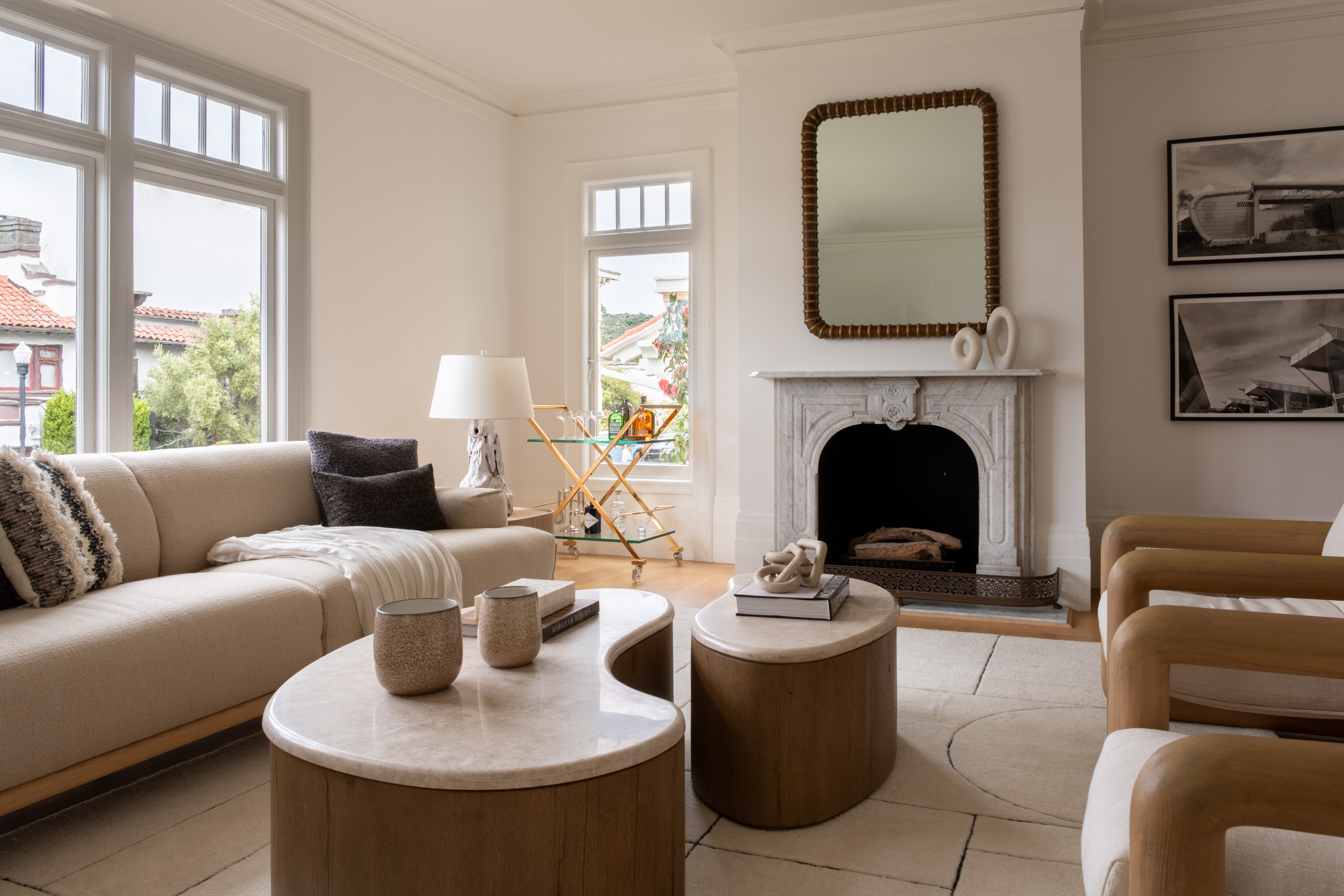 Bright Living Room With A Beige Sofa, Two Wooden Armchairs, Round Coffee Tables, A White Fireplace With A Mirror Above It, And Large Windows Letting In Natural Light. Decor Includes Neutral Tones And Minimalist Art.