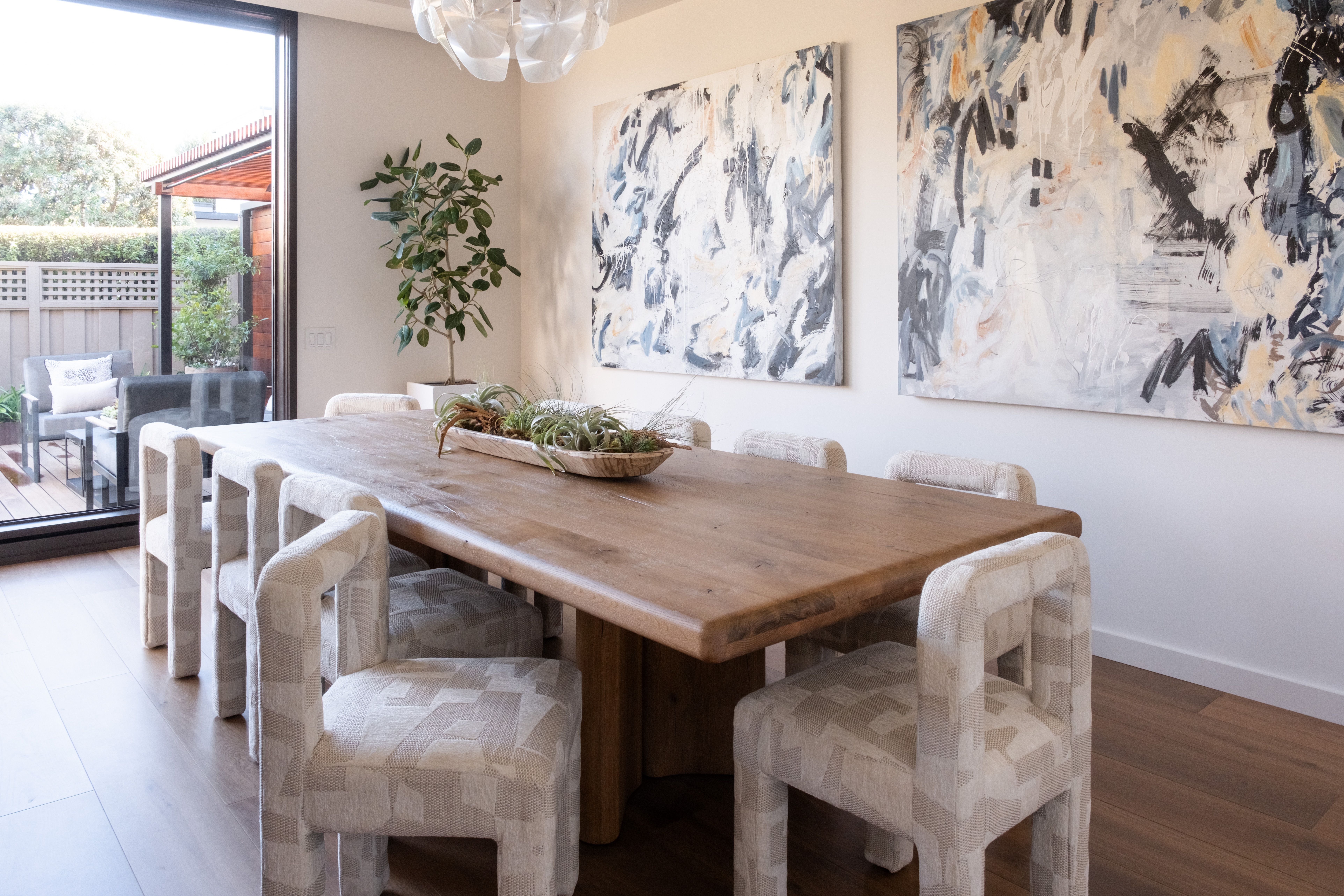Modern Dining Room With A Large Wooden Table, Eight Cushioned Chairs, Abstract Art On The Walls, A Plant In The Corner, And Large Windows Letting In Natural Light.