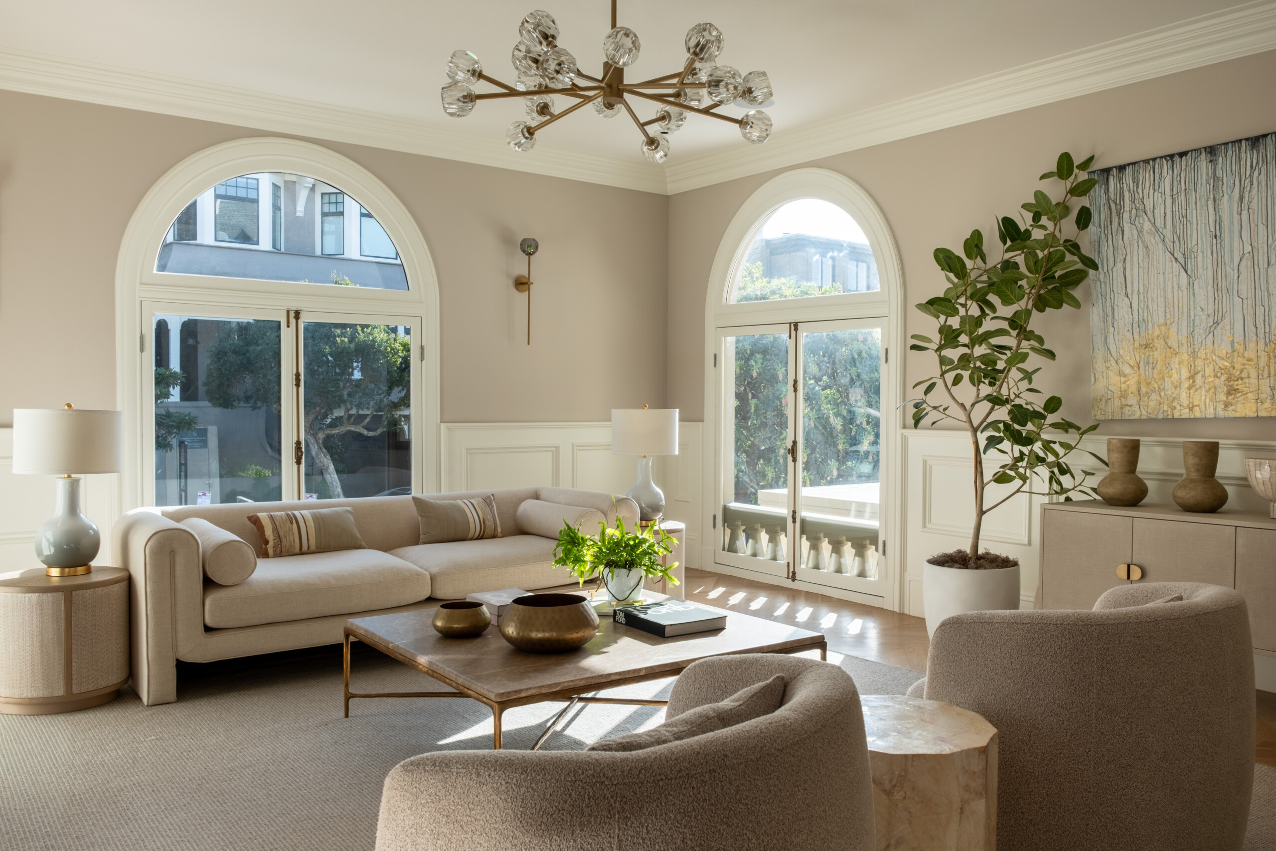 Bright Living Room With Arched Windows, Beige Walls, A Modern Chandelier, Cream Sofa, Two Armchairs, A Coffee Table, Large Plant, And Decorative Vases On A Console Under A Blue And Gold Abstract Painting.