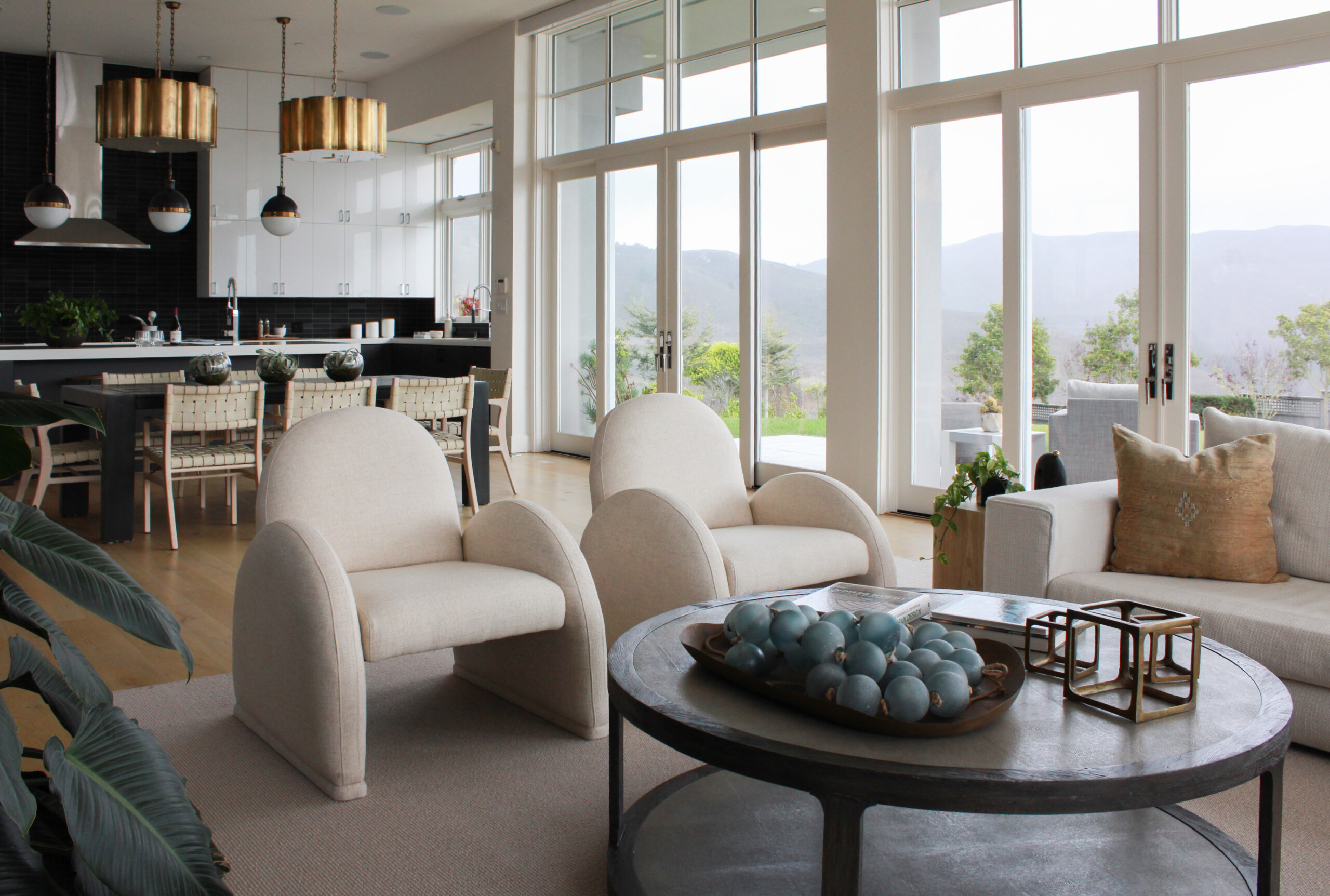 Modern Living Room With Cream Chairs, A Round Coffee Table With Blue Decorative Spheres, And Large Windows Showing A Scenic Outdoor View. Kitchen With Pendant Lights And A Dining Area Are Visible In The Background.