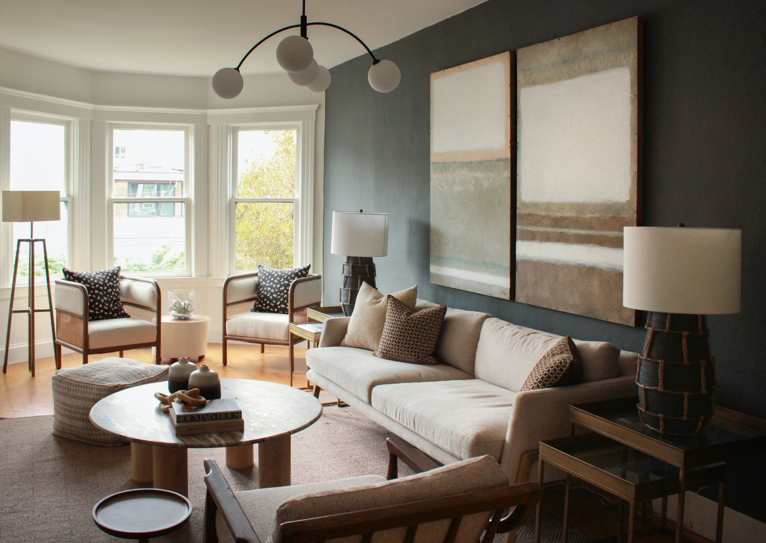 Modern Living Room With A Beige Sofa, Two Armchairs, Round Coffee Table, Large Abstract Wall Art, Floor And Table Lamps, And Bright Bay Windows Letting In Natural Light.