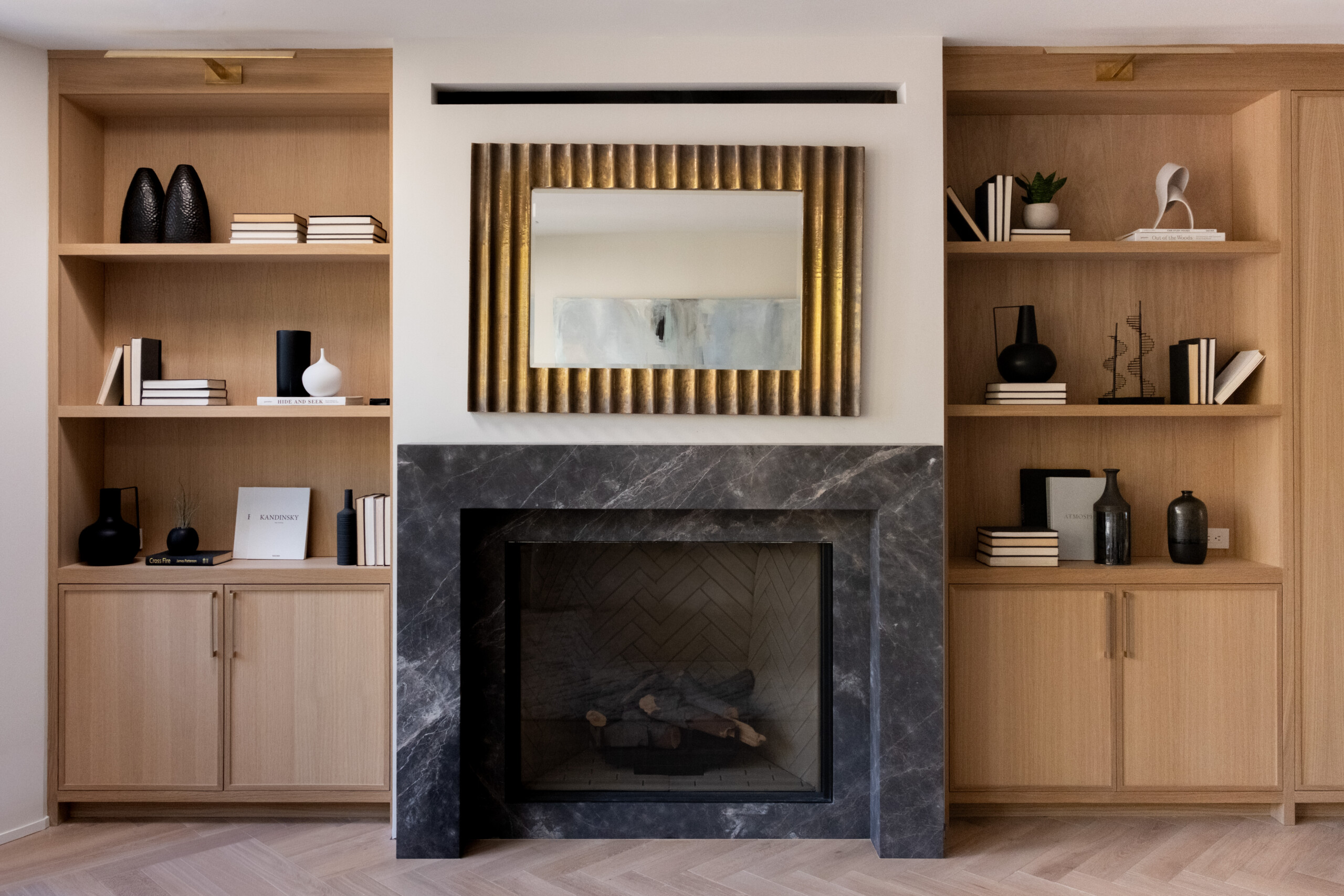 Modern Living Room With A Black Marble Fireplace, A Gold-Framed Mirror Above It, And Built-In Wooden Shelves Decorated With Books, Vases, And Minimalist Sculptures On Either Side.