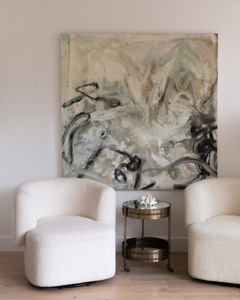 Two White Boucle Armchairs Sit Beside A Small Round Metal Table With A Decorative Item, In Front Of A Large Abstract Painting In Neutral Tones On A Light Wall.