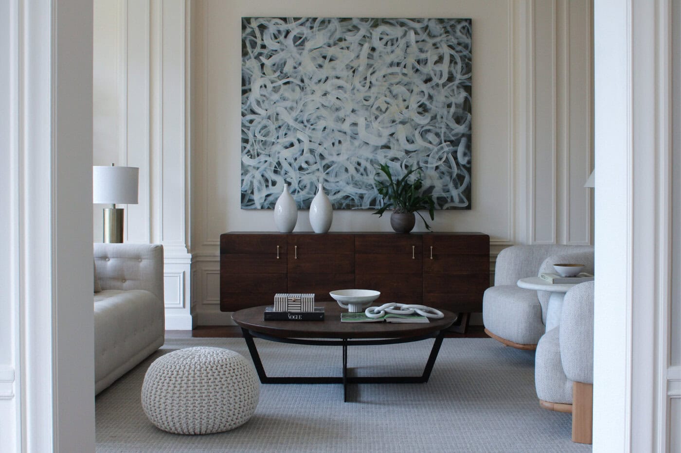 Modern Living Room With Neutral Furniture, A Dark Wood Sideboard, Abstract Art On The Wall, Round Coffee Table, Knit Pouf, And Decorative Vases And Books. The Room Has Soft Natural Light And Minimalist Decor.