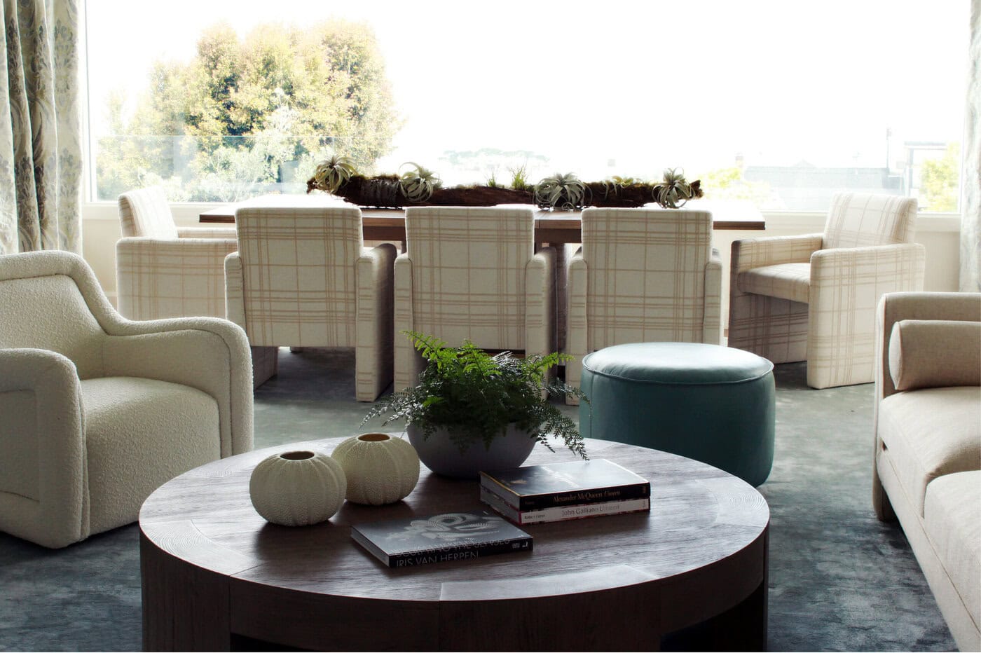A Modern Living Room With A Round Wooden Coffee Table, Two White Vases, Books, A Fern, Beige Armchairs, A Blue Ottoman, And A Dining Table With Plaid Chairs Near Large Windows Showing Trees Outside.