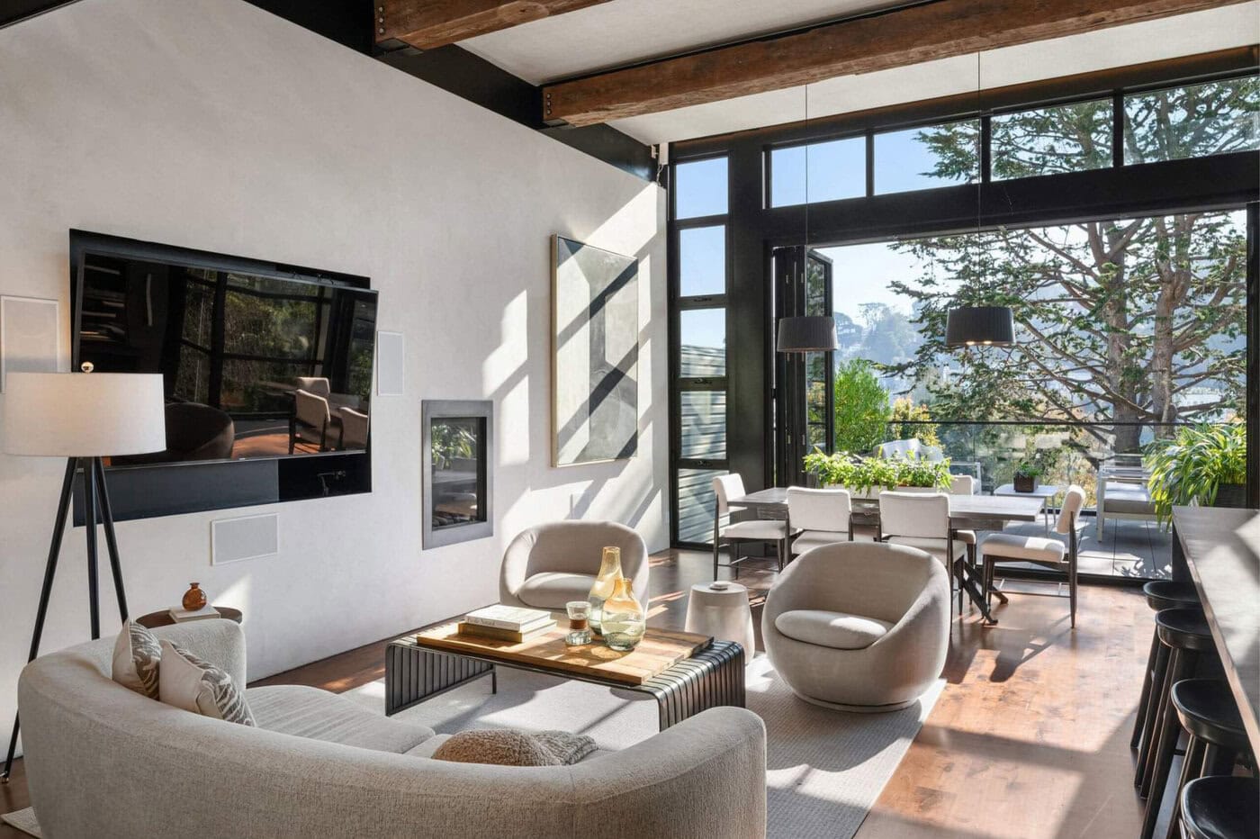 Modern Living And Dining Area With Large Windows, Neutral Furniture, Wood Beams, And Plenty Of Natural Light. There Are Trees And A View Of Mountains Visible Outside.