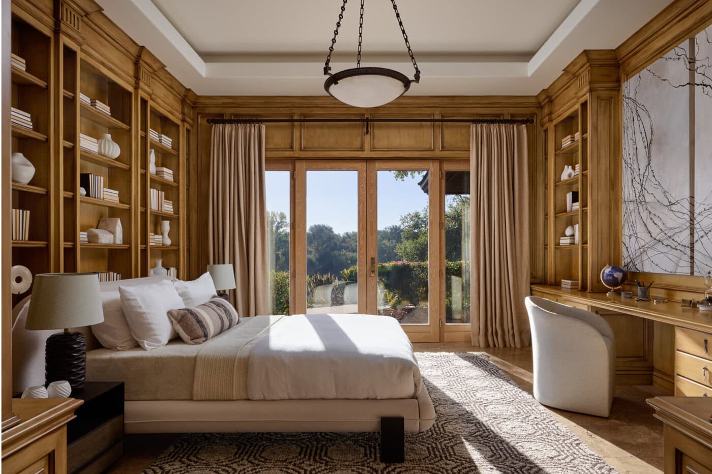 A Cozy Bedroom With Built-In Wooden Shelves, A Bed, Desk, Large Rug, And Glass Doors Opening To A Scenic Outdoor View, Letting In Natural Light. Decor Includes Books, Pottery, And A Large Abstract Wall Art.