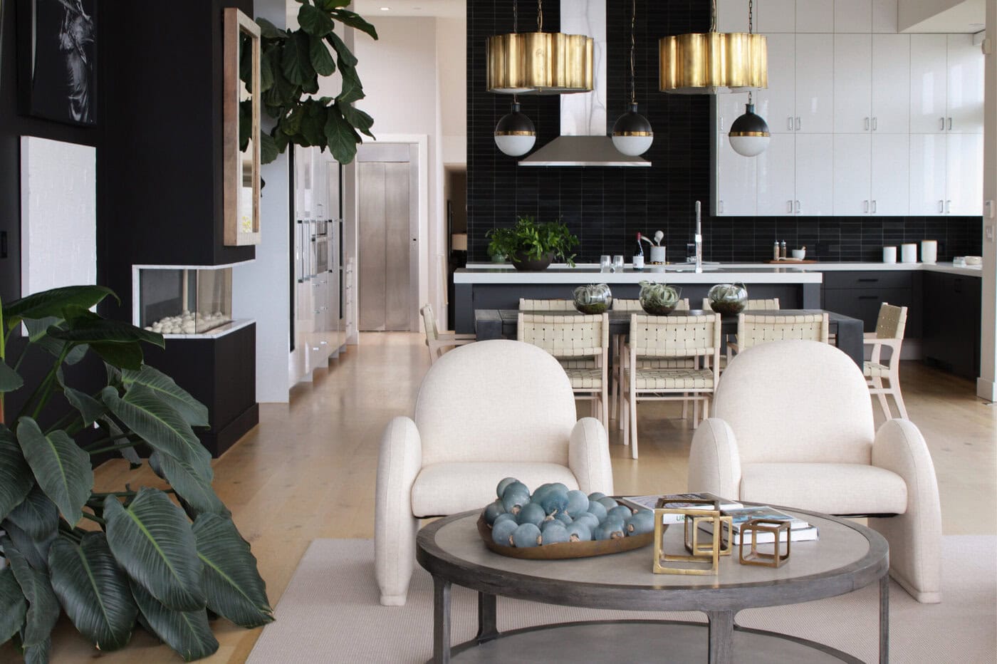 Modern Open-Concept Living Room And Kitchen With Neutral Furniture, Gold Light Fixtures, Large Indoor Plants, And A Round Coffee Table With Decorative Objects. White Cabinets And Black Accents Create A Sleek, Airy Feel.