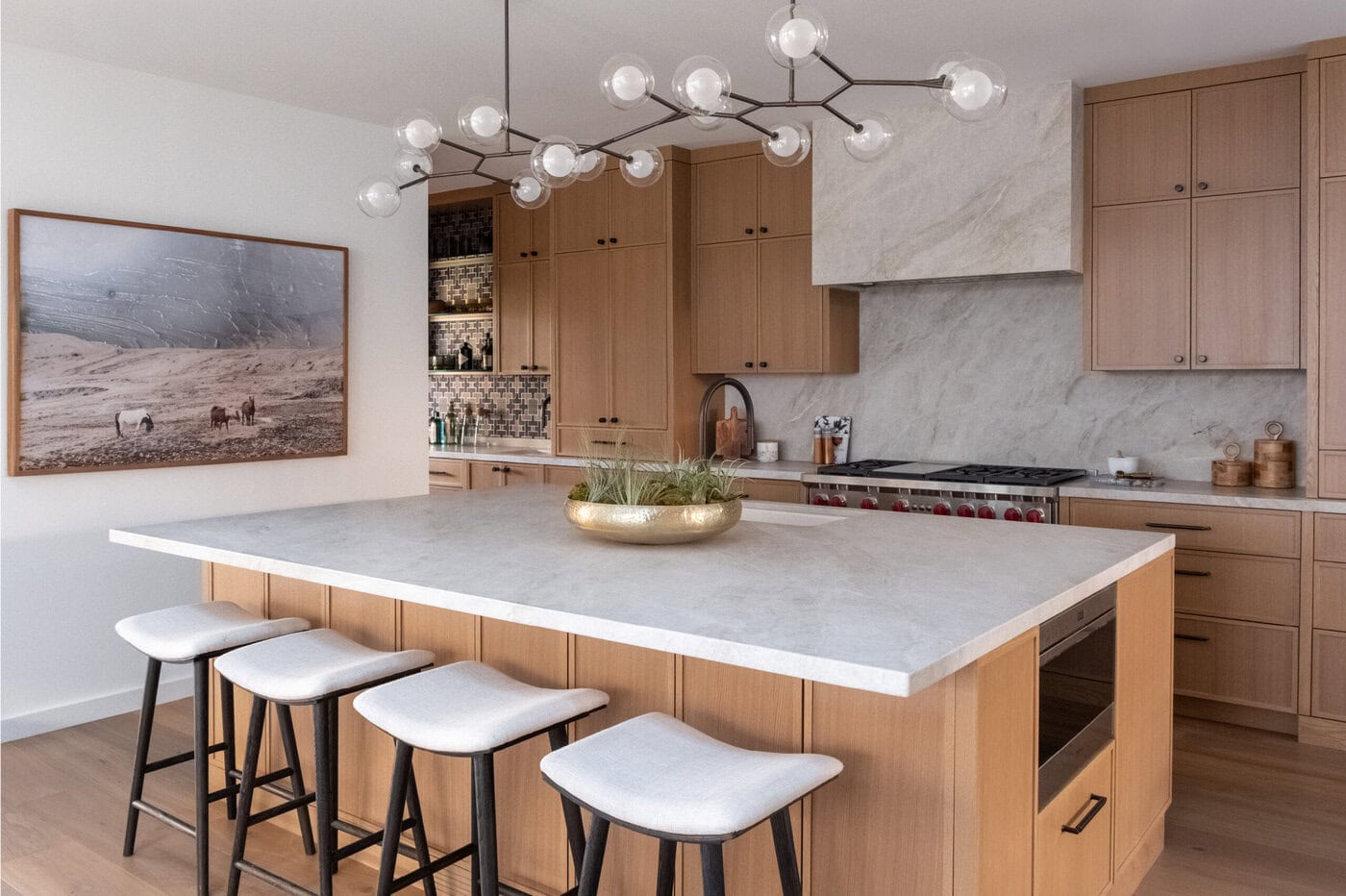 Modern Kitchen With Light Wood Cabinets, Marble Countertops, An Island With Four Cushioned Stools, A Plant Centerpiece, A Unique Chandelier, And A Large Wall Art Featuring Horses.