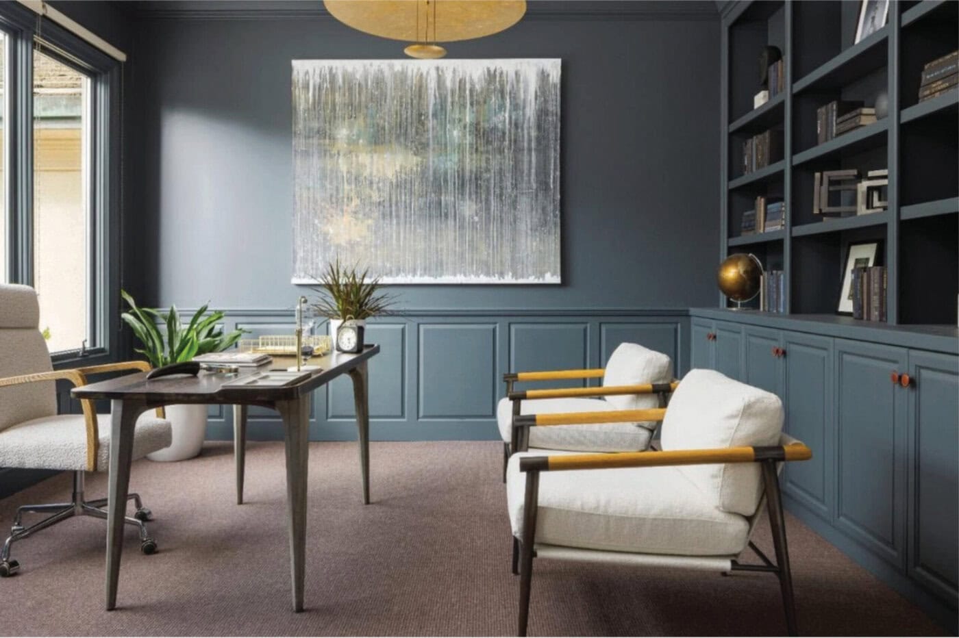 Modern Office With Gray Walls, Built-In Blue Shelves, Two White Armchairs, A Desk With Plants And Books, And Abstract Artwork On The Wall. Natural Light Comes In Through A Large Window On The Left.