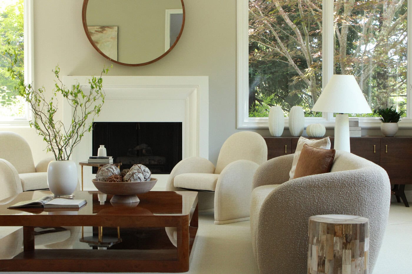 Modern Living Room With Cream-Colored Chairs, A Wooden Coffee Table, A Fireplace, Green Branches In A Vase, Decorative Objects, Large Windows, And A Round Mirror Above The Mantel. Natural Light Fills The Space.