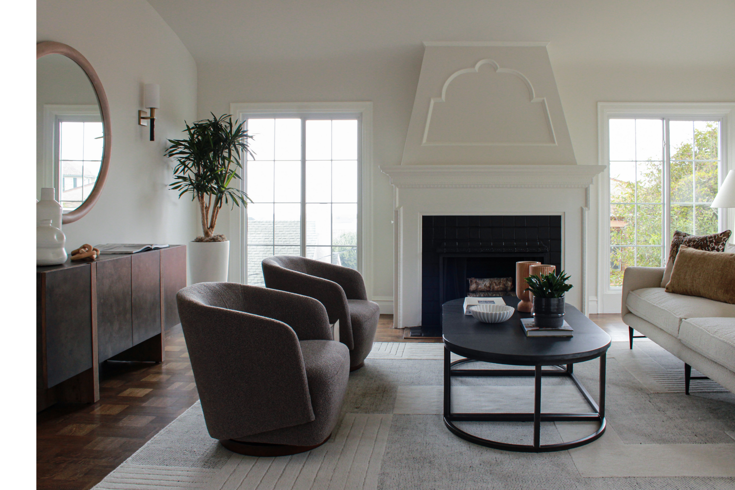 Modern Living Room With Two Gray Chairs, A Beige Sofa, Black Coffee Table, Potted Plants, Sideboard, And A White Fireplace. Large Windows Let In Natural Light, Creating A Bright, Inviting Space.