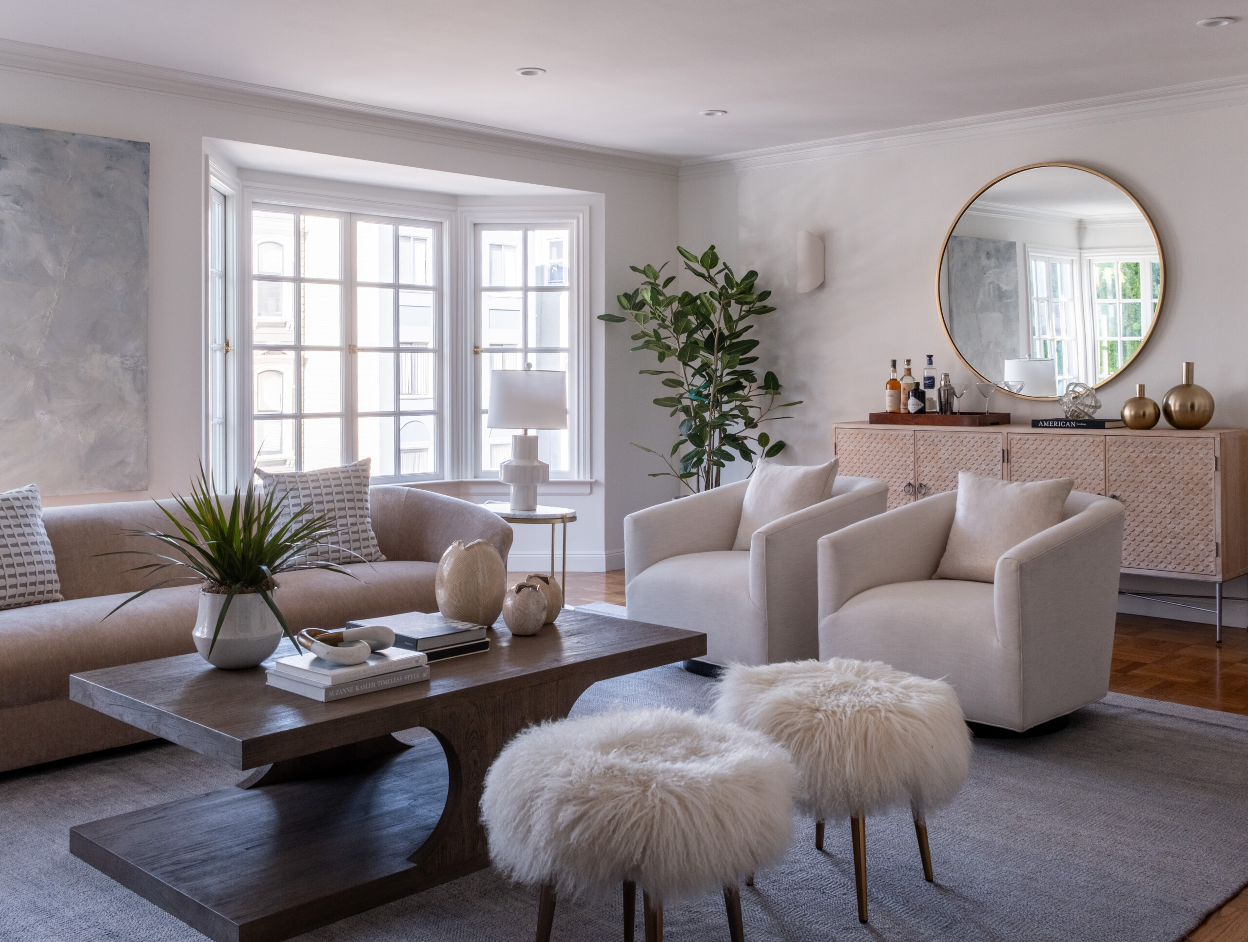 Modern Living Room With Beige Sofa, Two White Armchairs, A Wooden Coffee Table, Two Fuzzy Stools, A Large Plant, And A Round Mirror Above A Console Table With Decorative Vases. Light Streams In From Large Windows.