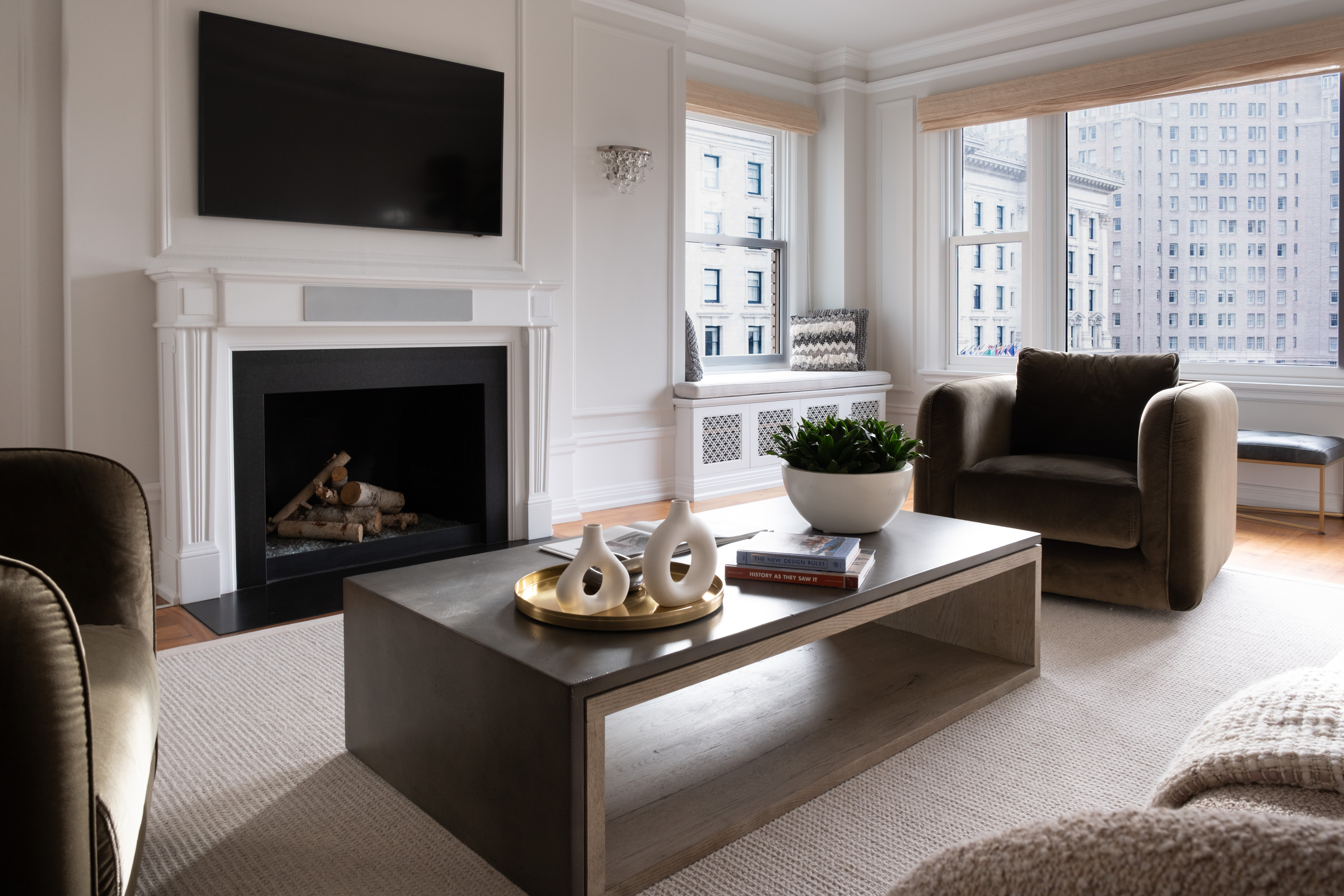 Modern Living Room With A Fireplace, Wall-Mounted Tv, Two Armchairs, A Coffee Table With Decor Items, A Potted Plant, Large Windows, And A Cityscape View. Neutral Colors Create A Calm, Inviting Atmosphere.